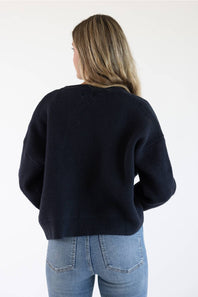 Back view of a female model wearing the Things Between Alyssa Cardigan in Navy, highlighting the drop-shoulder seam detail, relaxed fit, and ribbed hemline.