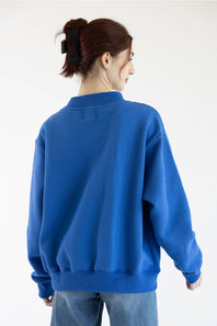 A woman with her hair in a high ponytail is shown from the back wearing a vibrant royal blue crewneck sweatshirt and light wash blue jeans against a plain white studio background.