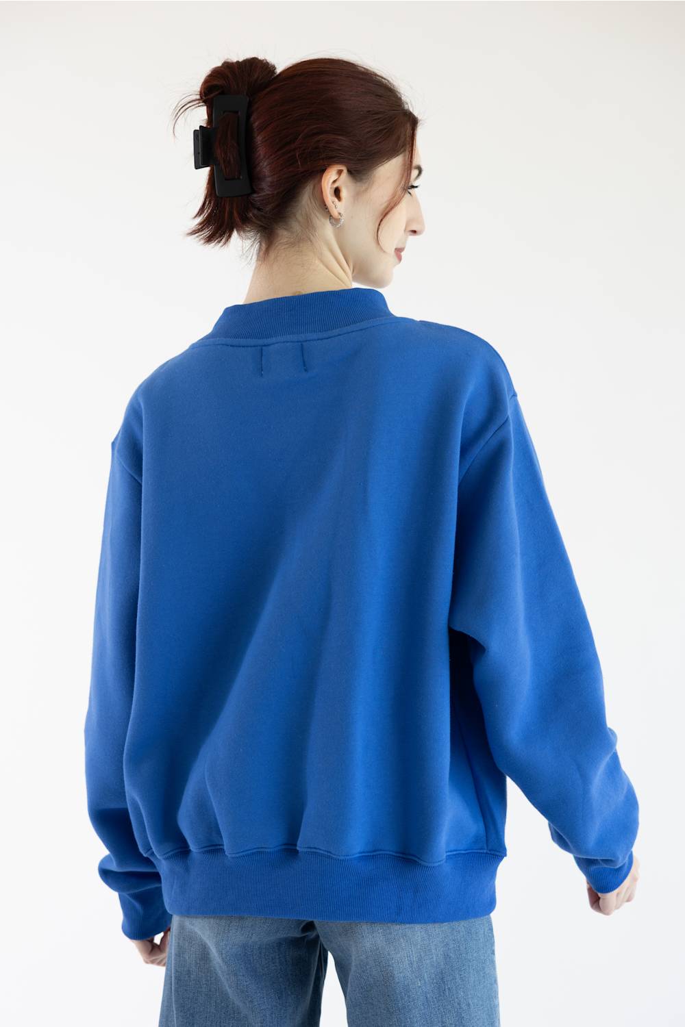 A woman with her hair in a high ponytail is shown from the back wearing a vibrant royal blue crewneck sweatshirt and light wash blue jeans against a plain white studio background.