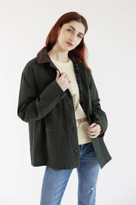 A woman with red hair wearing an olive green utility jacket with a brown corduroy collar, layered over a cream sweater and blue jeans, photographed against a plain white studio background.