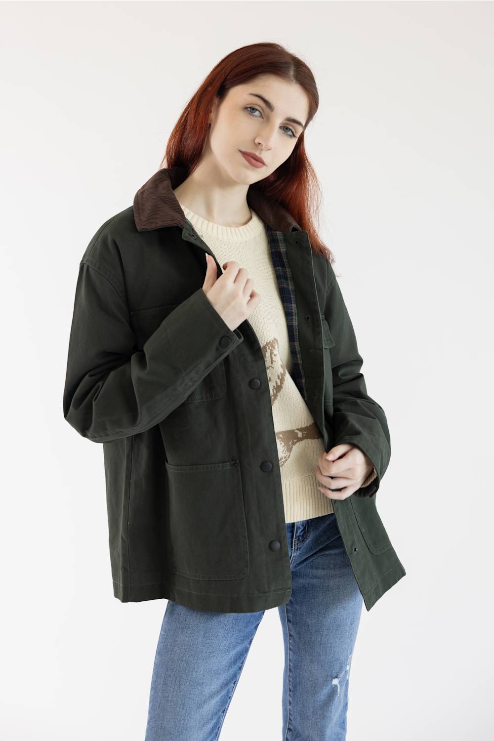 A woman with red hair wearing an olive green utility jacket with a brown corduroy collar, layered over a cream sweater and blue jeans, photographed against a plain white studio background.
