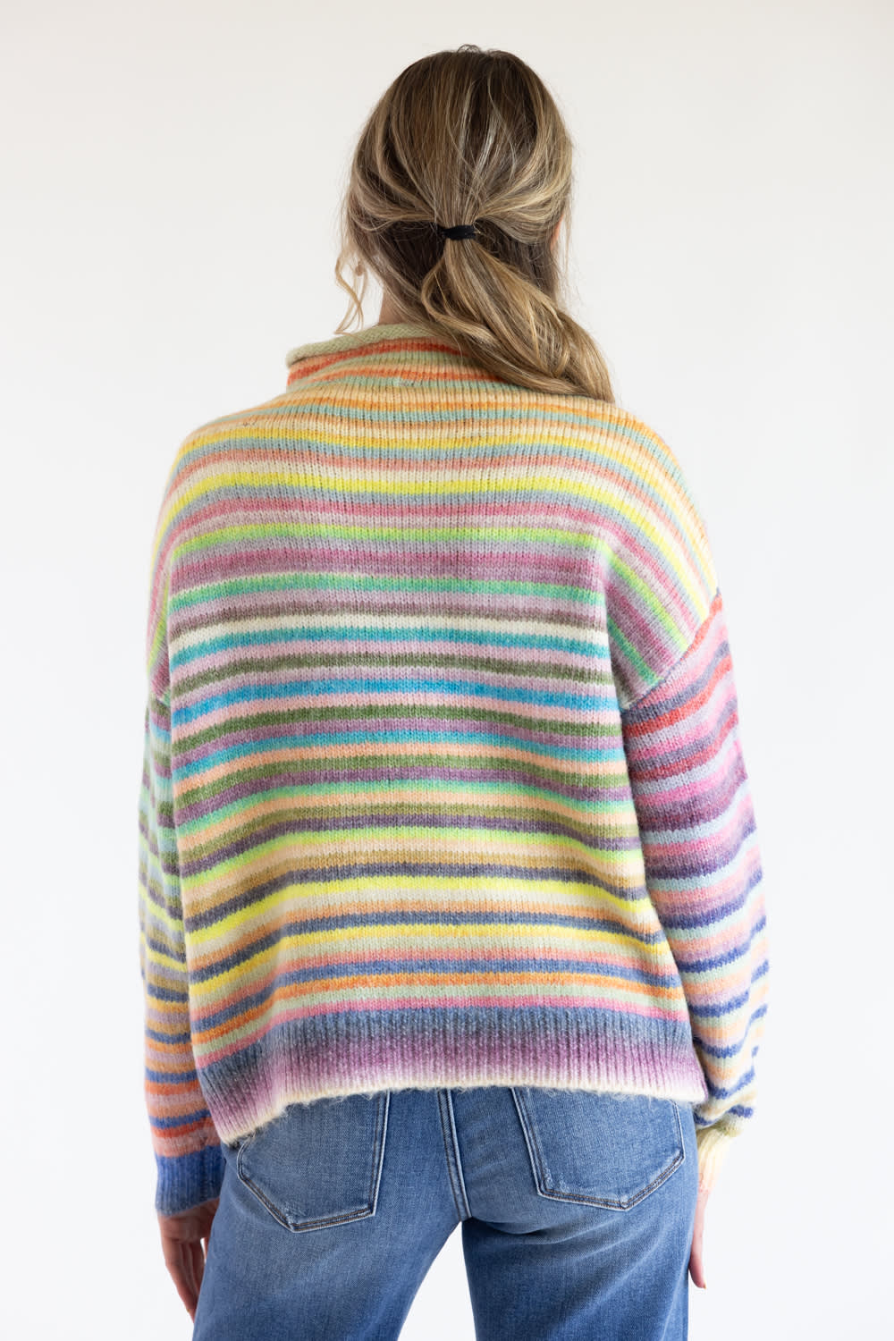 A woman viewed from the back wearing a rainbow colored striped sweater with long sleeves and blue denim jeans standing in front of a white studio background