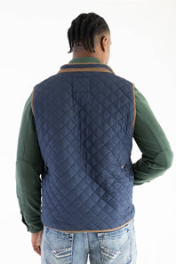 A man viewed from behind wearing a navy blue diamond-quilted puffer vest with tan trim over a green long-sleeve shirt and blue jeans, against a plain white background.