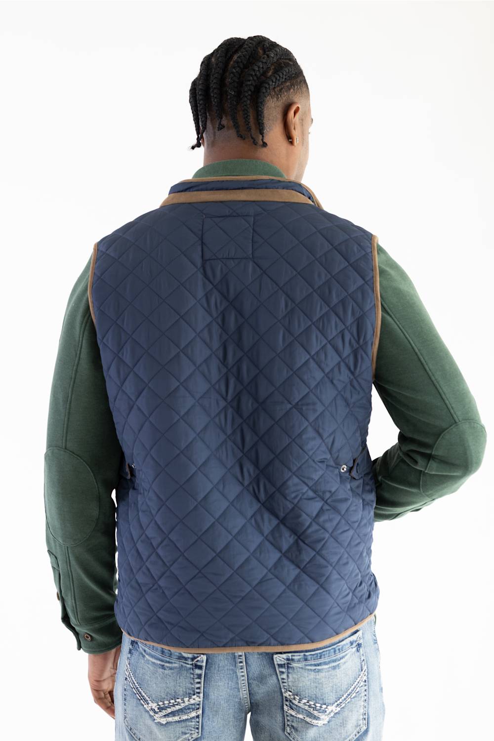 A man viewed from behind wearing a navy blue diamond-quilted puffer vest with tan trim over a green long-sleeve shirt and blue jeans, against a plain white background.