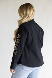 A woman shown from the back wearing a black jacket with 'ARIAT' printed vertically in gold lettering down the left sleeve, paired with light blue jeans against a plain light gray background.