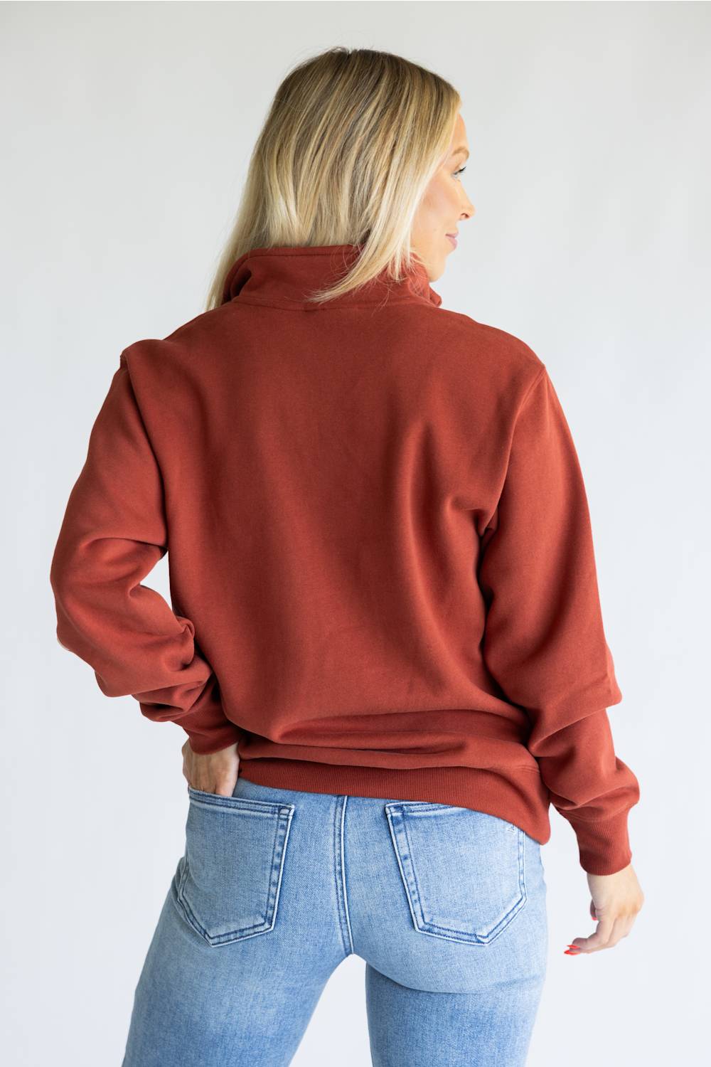 A woman shown from the back wearing a rust-colored long-sleeve mock neck sweatshirt and light blue jeans, photographed against a plain white studio background.