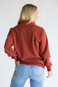 A woman shown from the back wearing a rust-colored long-sleeve mock neck sweatshirt and light blue jeans, photographed against a plain white studio background.