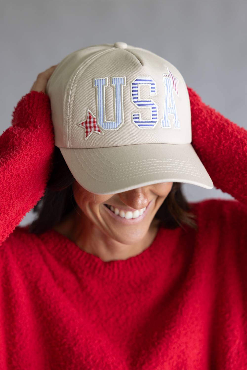 Woman wearing a beige cap with 'USA' and a checkered star design, smiling against a gray background.