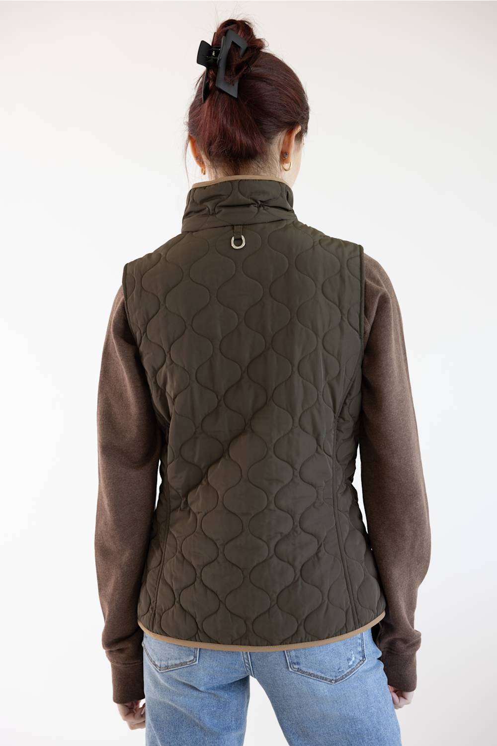 A woman viewed from the back wearing an olive green quilted vest over a brown long-sleeve shirt and blue jeans, against a plain white background.
