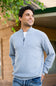 A man wearing a light blue quarter-zip knit sweater over a white undershirt and blue jeans, standing outdoors in front of a house with wood siding and green foliage.