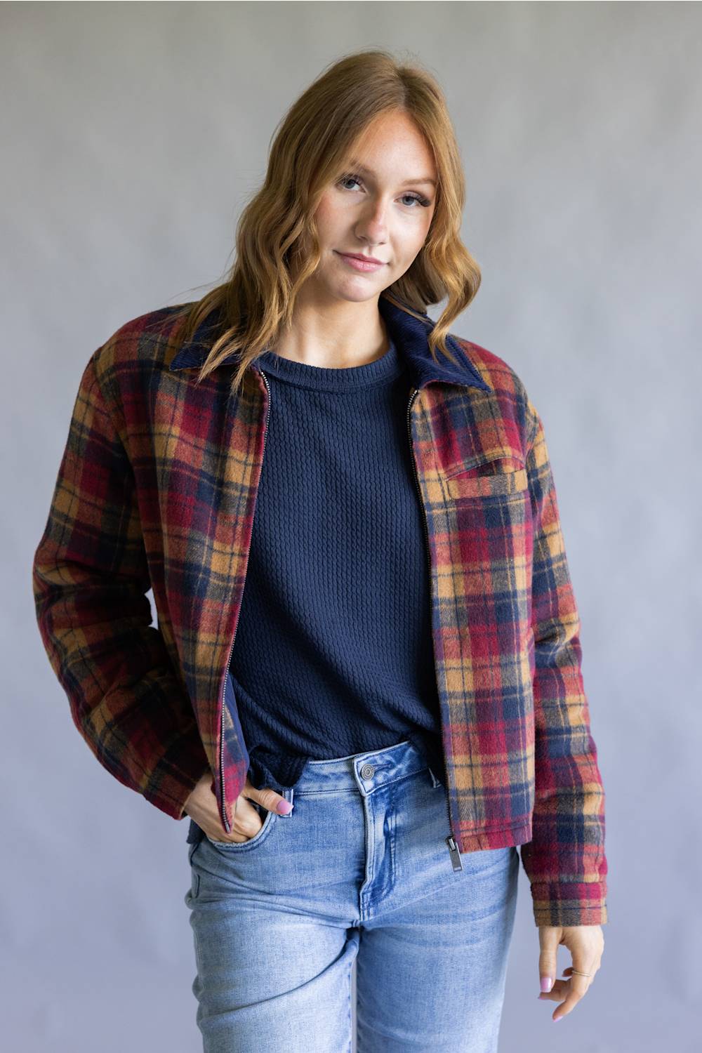A woman wearing a red, navy, and yellow plaid flannel overshirt over a navy ribbed knit top and light blue jeans, standing against a neutral gray studio background.