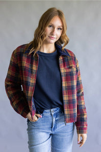 A woman wearing a red, navy, and yellow plaid flannel overshirt over a navy ribbed knit top and light blue jeans, standing against a neutral gray studio background.