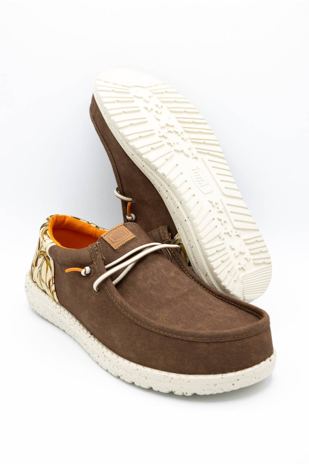 A pair of brown suede slip-on shoes with white speckled soles, white laces, and a gold patterned heel detail, shown on a white background with one shoe standing upright to display the sole.