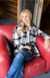 A woman with blonde hair sits in a red leather armchair, wearing a black, white, and gray plaid flannel shirt over a white top and blue jeans, holding a pair of binoculars.