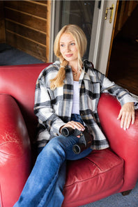 A woman with blonde hair sits in a red leather armchair, wearing a black, white, and gray plaid flannel shirt over a white top and blue jeans, holding a pair of binoculars.