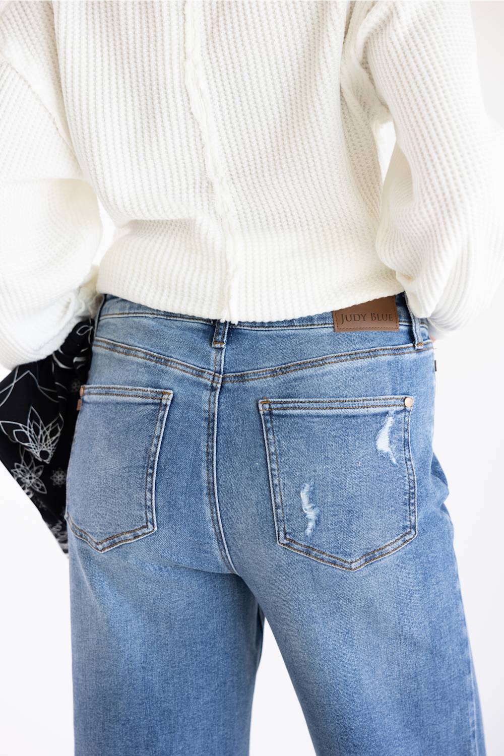 Close-up back view of a person wearing light blue distressed jeans with a white ribbed knit sweater and a black patterned bandana tucked into one pocket.
