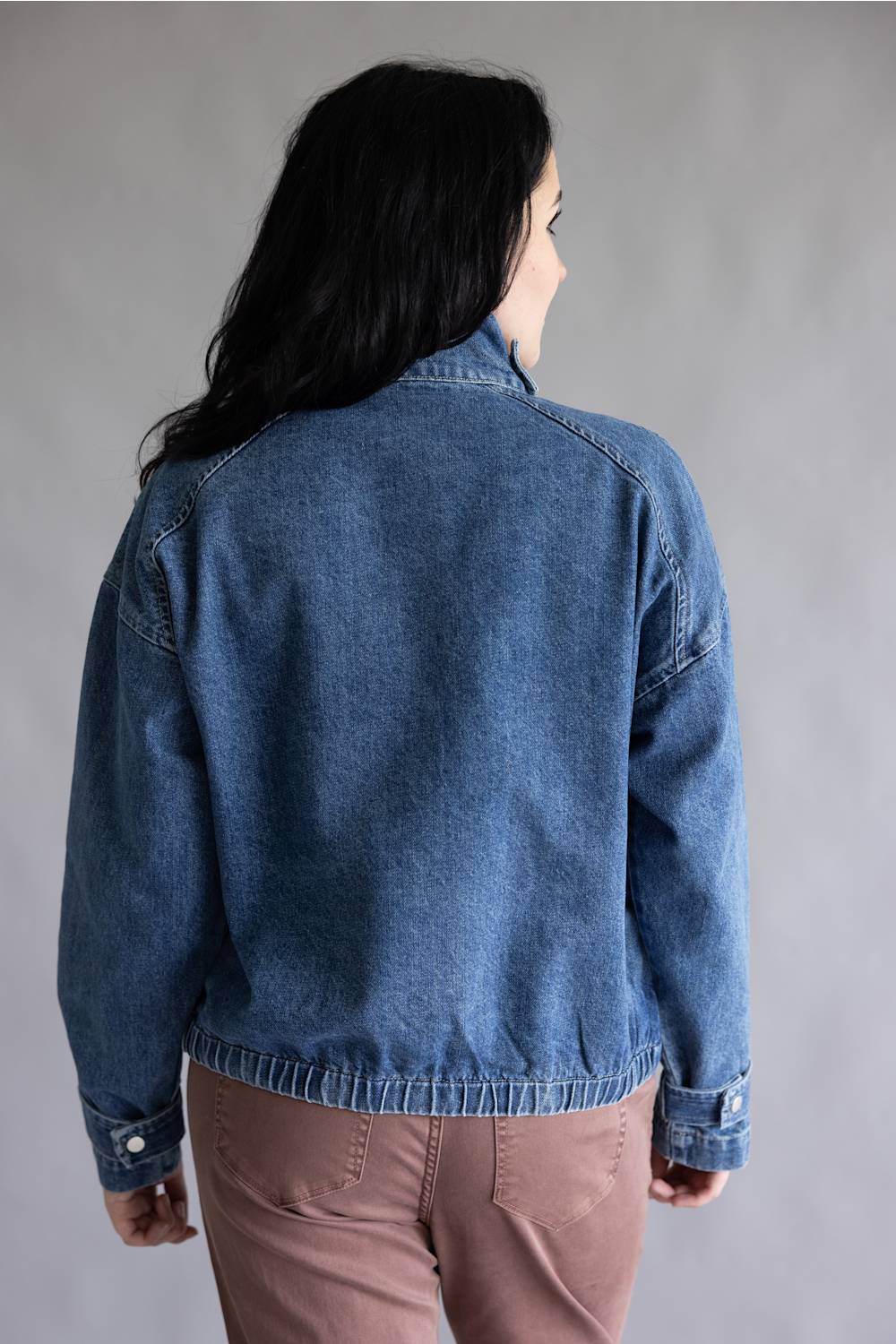 A woman viewed from the back wearing a medium wash blue denim jacket and dusty rose pants, standing against a neutral gray studio background.