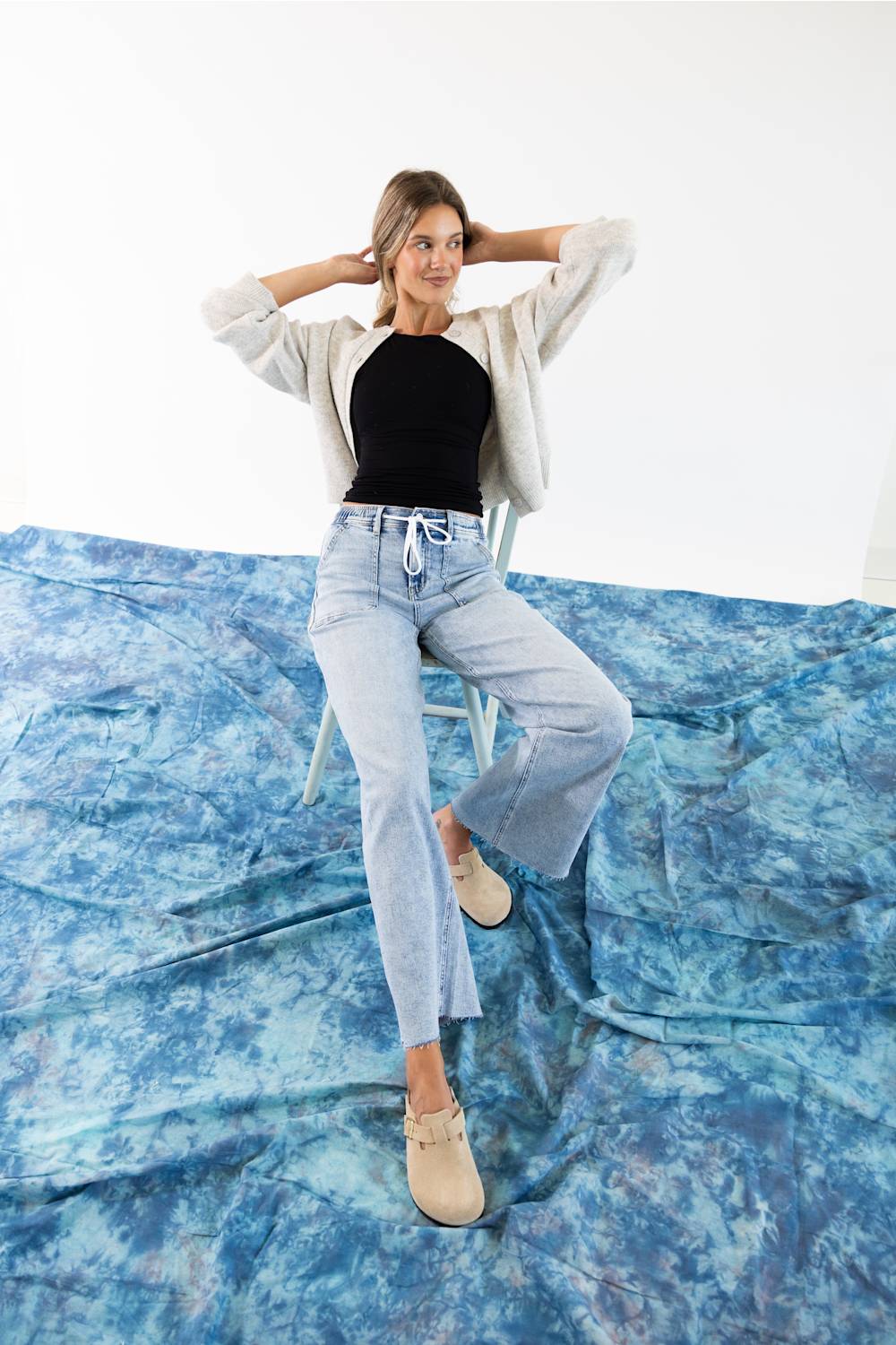 A woman modeling a light gray knit cardigan with a button-up front and drawstring detail, paired with light blue jeans against a plain white background.