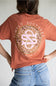 A person wearing a rust-orange t-shirt from the back, featuring a large circular graphic with the text 'SIMPLY SOUTHERN' and a pink-and-white monogram. The person is also wearing blue jeans against a plain light gray background.