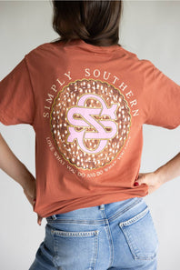 A person wearing a rust-orange t-shirt from the back, featuring a large circular graphic with the text 'SIMPLY SOUTHERN' and a pink-and-white monogram. The person is also wearing blue jeans against a plain light gray background.