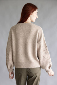 A woman with long auburn hair is shown from the back wearing a beige knit sweater with floral embroidery on the sleeves, paired with olive green pants, against a neutral gray studio background.