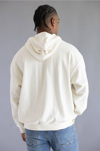 A man viewed from behind wearing an oversized cream-colored hoodie and blue jeans, standing against a solid gray studio background.
