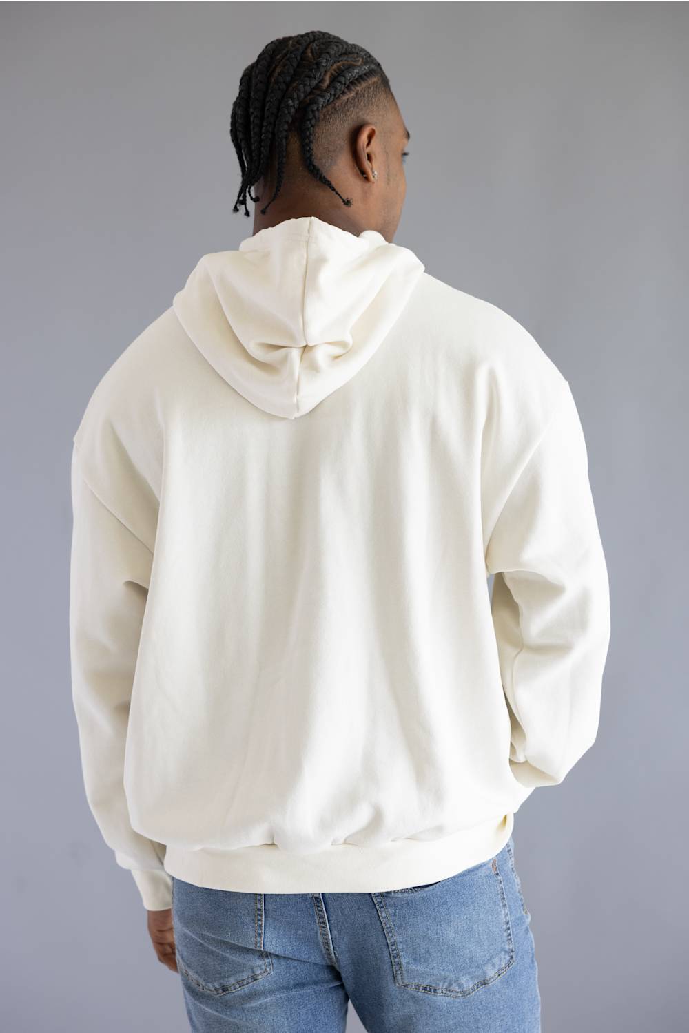 A man viewed from behind wearing an oversized cream-colored hoodie and blue jeans, standing against a solid gray studio background.