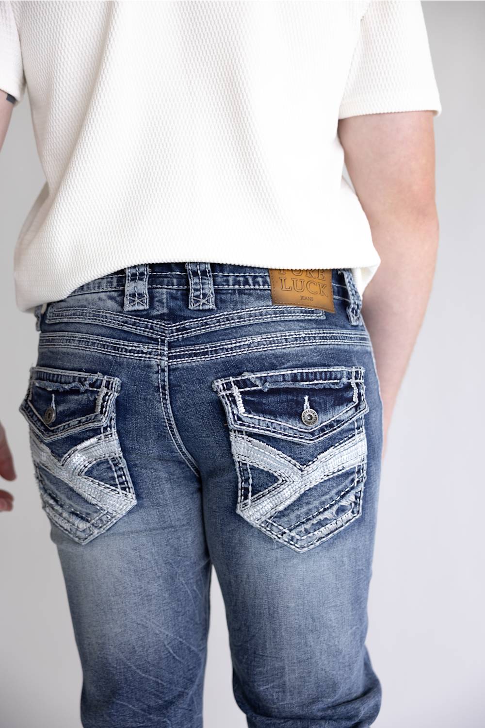 Rear view of a person wearing blue denim jeans with decorative distressed stitching and patch pockets, paired with a white ribbed short-sleeve top against a light gray background.