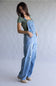 A woman modeling a pair of light blue denim overall with distressed details, worn over a sage green t-shirt and beige shoes, against a neutral gray studio background.