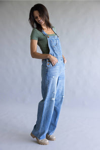 A woman modeling a pair of light blue denim overall with distressed details, worn over a sage green t-shirt and beige shoes, against a neutral gray studio background.