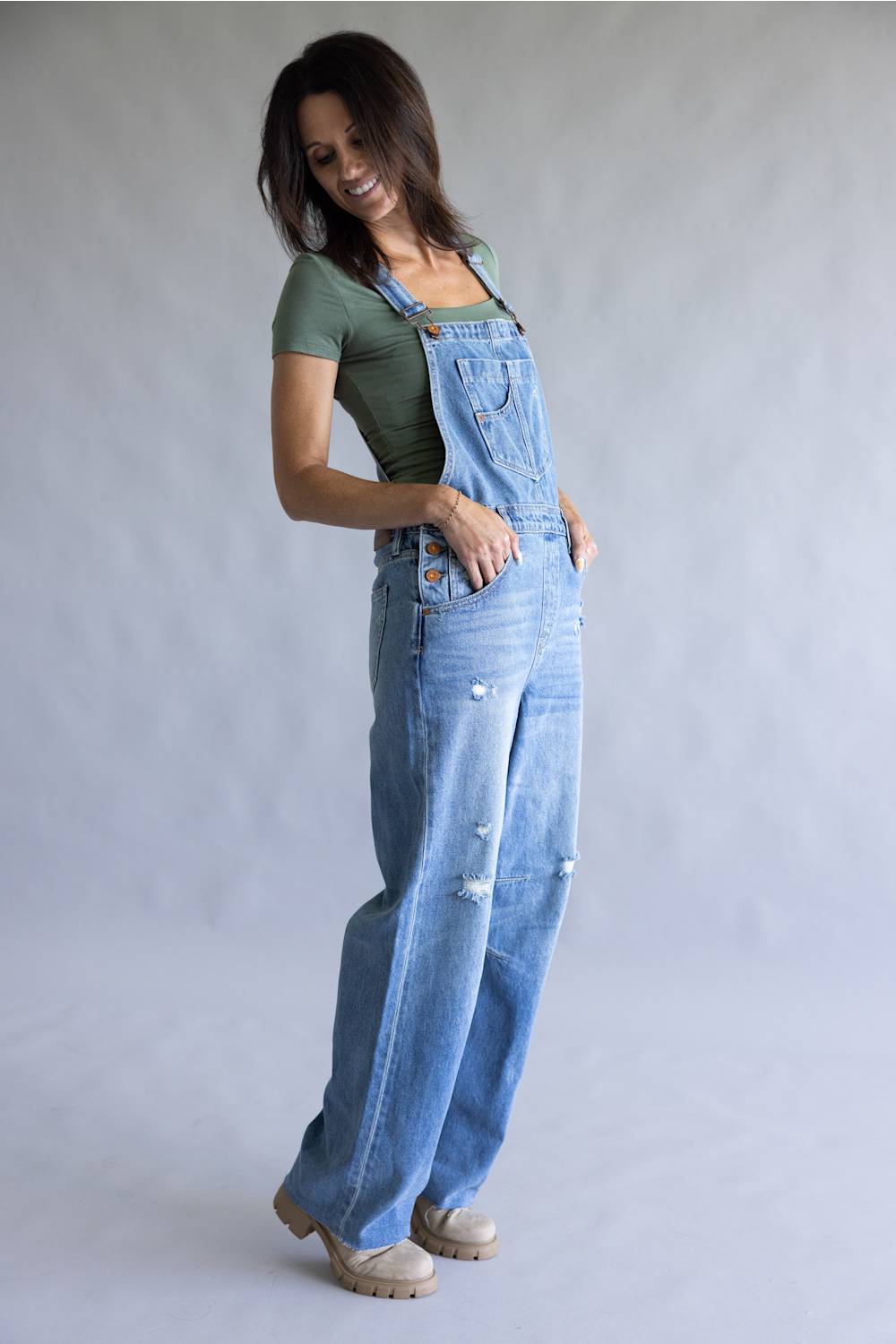 A woman modeling a pair of light blue denim overall with distressed details, worn over a sage green t-shirt and beige shoes, against a neutral gray studio background.