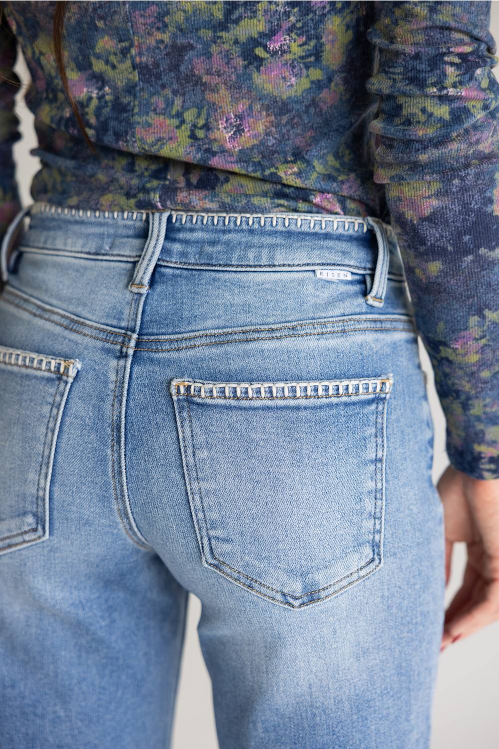 Close-up back view of a person wearing light-wash blue denim jeans with decorative contrast stitching around the back pockets, paired with a floral long-sleeve top.