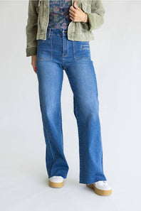 A person wearing blue wide-leg jeans with a faded wash, styled with an olive green jacket, a floral top, and white platform sneakers against a plain light gray studio background.