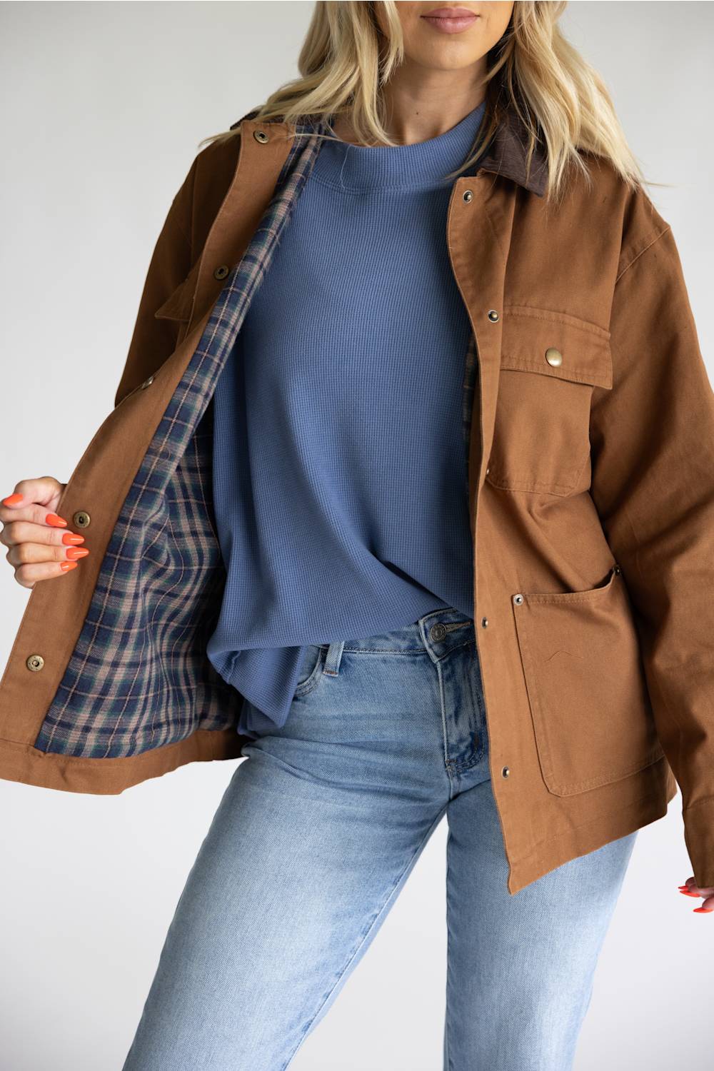 A woman wearing a brown utility jacket with a blue plaid lining, layered over a slate blue top and light wash jeans.