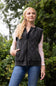 A woman wearing a dark gray diamond-quilted zip-up vest over a light pink long-sleeve shirt and blue jeans, standing outdoors against a blurred green foliage background.