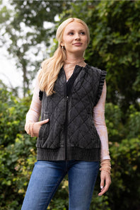 A woman wearing a dark gray diamond-quilted zip-up vest over a light pink long-sleeve shirt and blue jeans, standing outdoors against a blurred green foliage background.