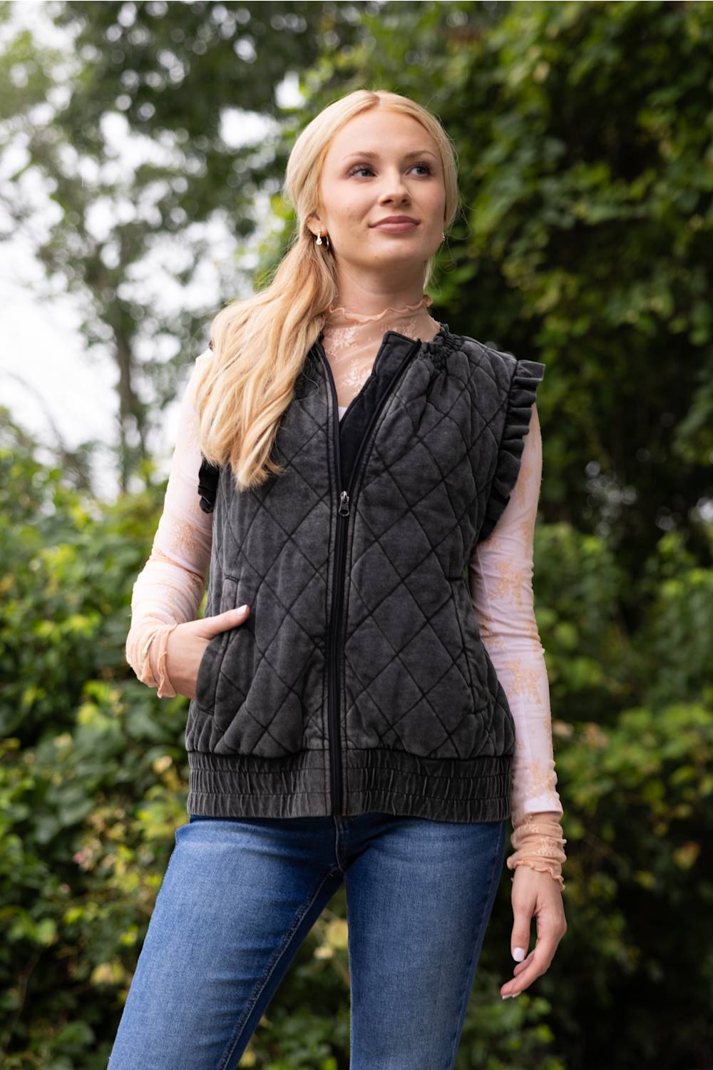 A woman wearing a dark gray diamond-quilted zip-up vest over a light pink long-sleeve shirt and blue jeans, standing outdoors against a blurred green foliage background.