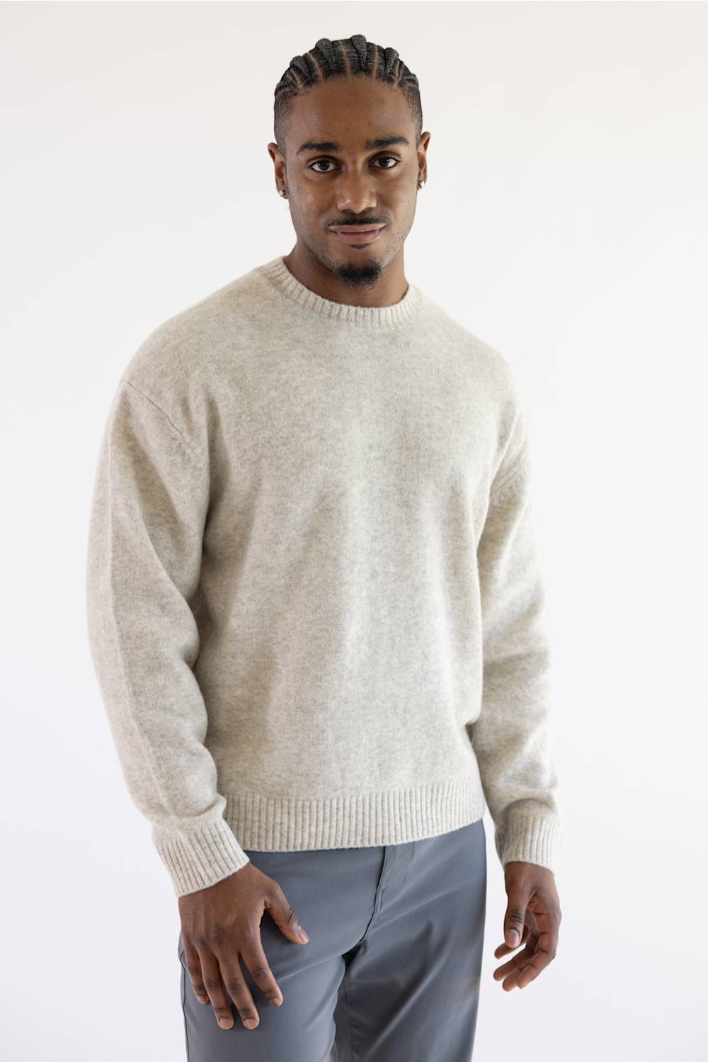 A man wearing a light beige knit crewneck sweater and gray trousers, standing against a plain white studio background.
