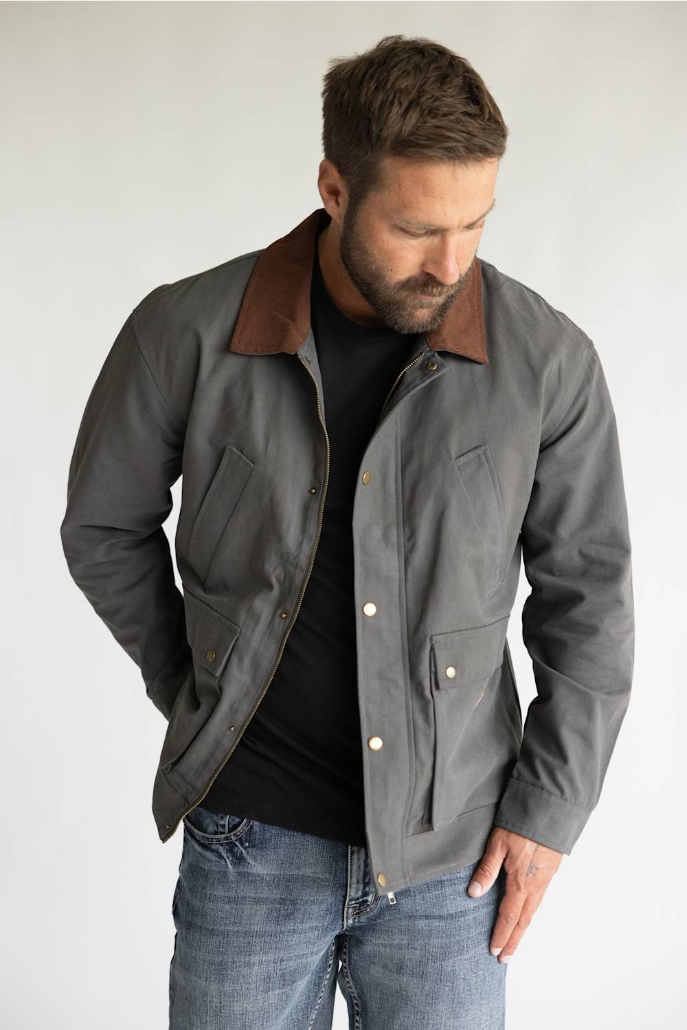 A man wearing a gray waxed cotton utility jacket with a brown corduroy collar, styled over a black t-shirt and blue jeans, standing against a light gray studio background.