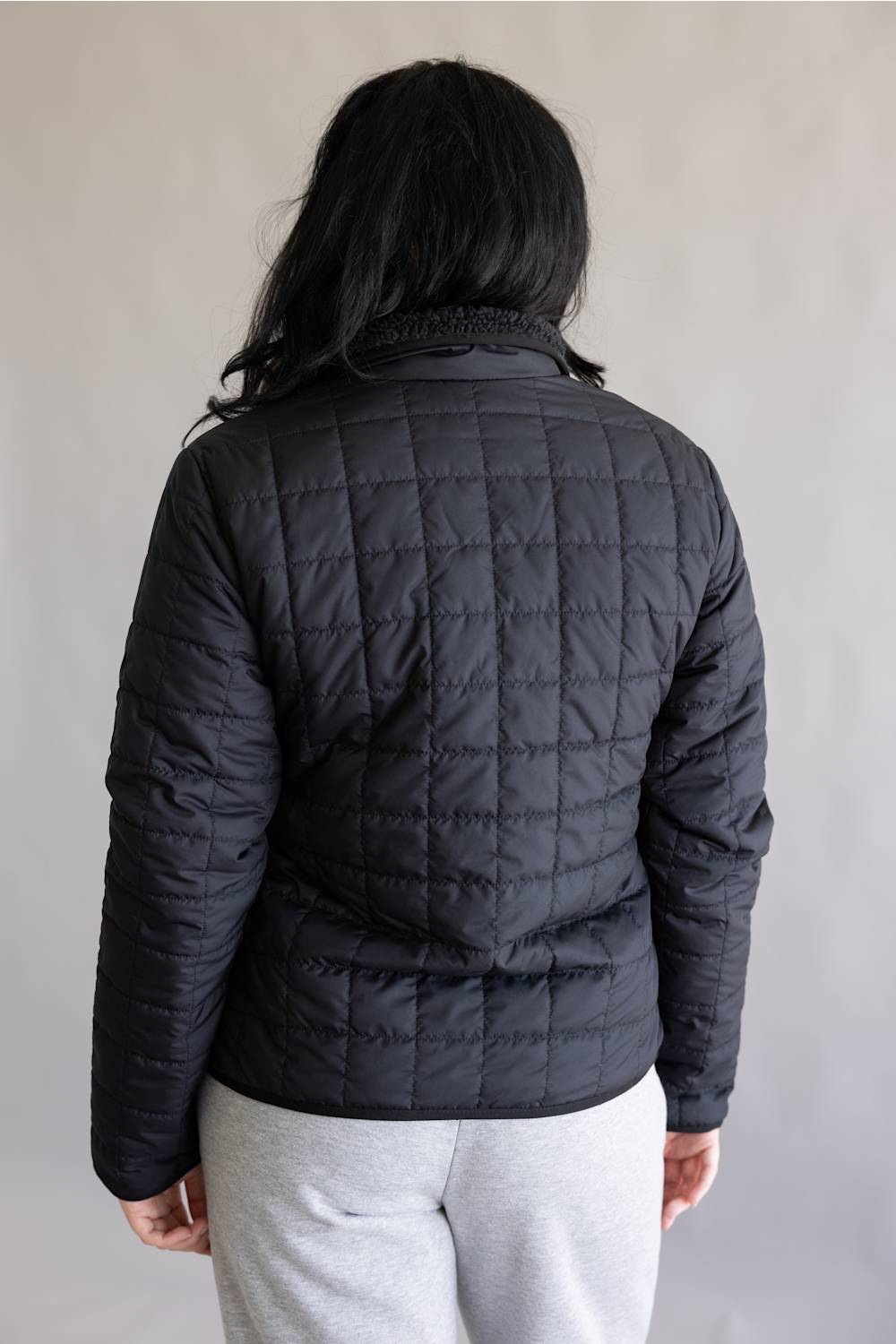 A person shown from the back wearing a black quilted puffer jacket and light gray sweatpants against a plain light gray background.