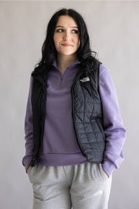 A woman wearing a black quilted vest over a purple quarter-zip sweatshirt and gray sweatpants, standing against a plain light gray background. The vest has a white The North Face logo on the chest.