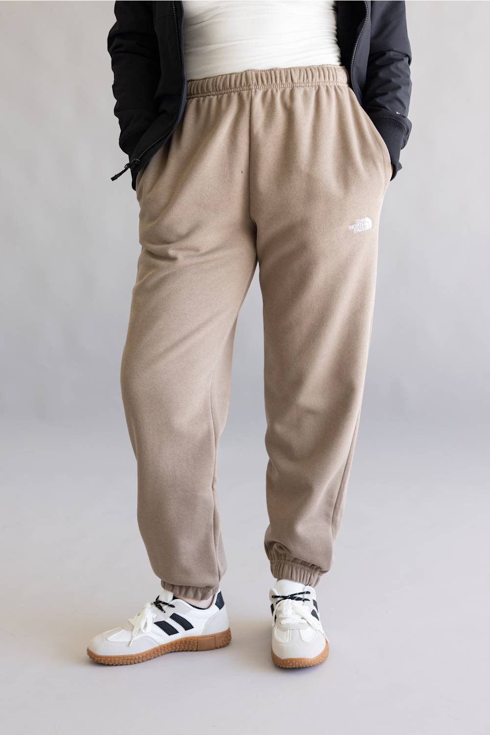 A person wearing beige jogger sweatpants with an elastic waistband and cuffs, styled with a white top, black jacket, and white sneakers with black stripes, shown from the waist down against a light gray studio background.