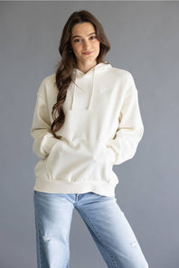 A woman wearing an oversized cream-colored hoodie with drawstrings and a kangaroo pocket, paired with light-wash blue jeans, standing against a solid gray studio background.