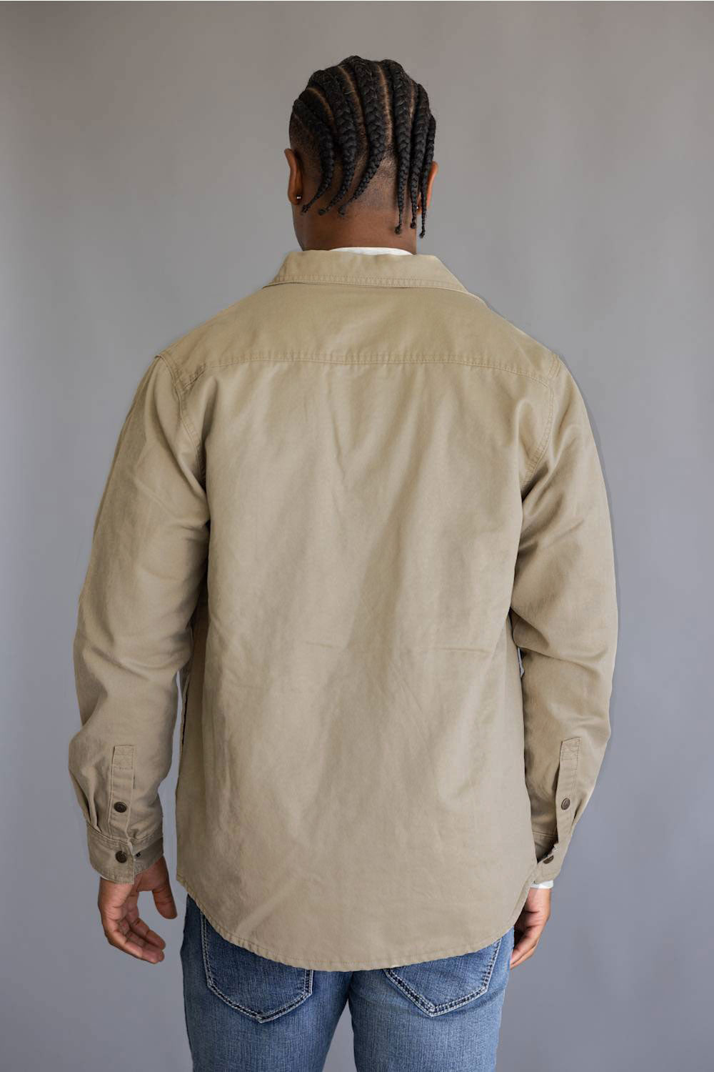 A man viewed from behind wearing a light tan long-sleeve button-up shirt and blue jeans, standing against a plain gray studio background.