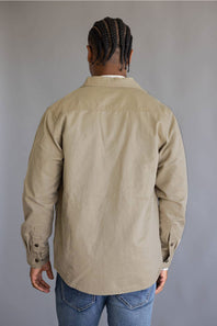 A man viewed from behind wearing a light tan long-sleeve button-up shirt and blue jeans, standing against a plain gray studio background.