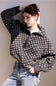 A woman wearing a black and white geometric patterned cropped jacket with a half-zip front, paired with light blue jeans, posing against a neutral beige wall.