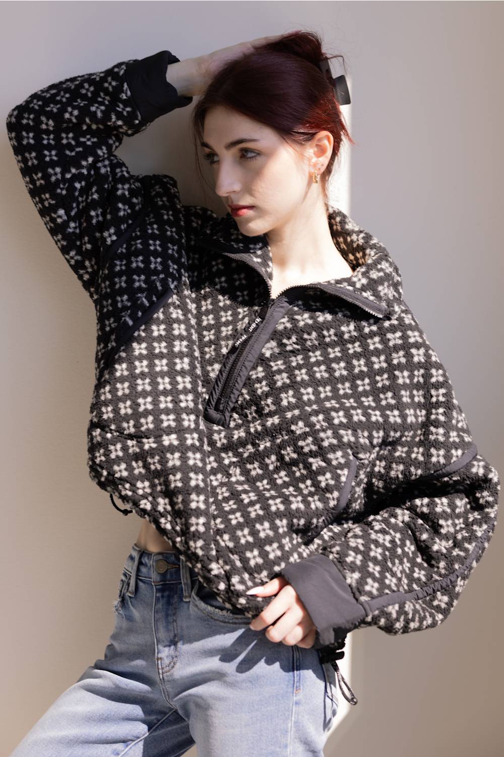 A woman wearing a black and white geometric patterned cropped jacket with a half-zip front, paired with light blue jeans, posing against a neutral beige wall.