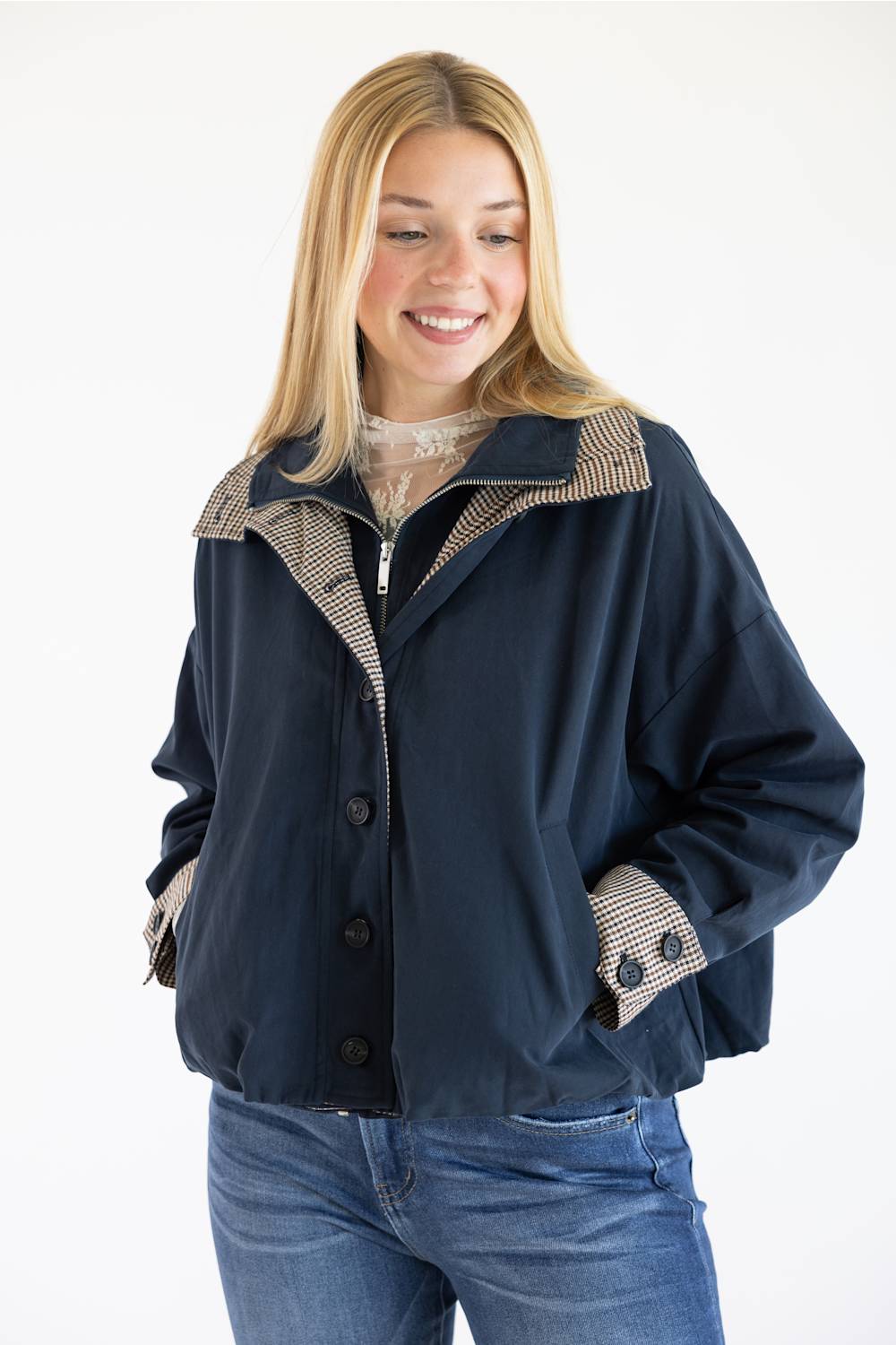 A smiling blonde woman wearing a navy blue jacket with beige and black patterned lining details on the collar, cuffs, and pockets, paired with blue jeans against a plain white background.