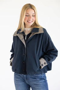 A smiling blonde woman wearing a navy blue jacket with beige and black patterned lining details on the collar, cuffs, and pockets, paired with blue jeans against a plain white background.