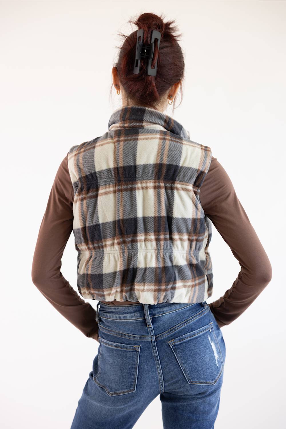 A woman shown from behind wearing a brown long-sleeve shirt under a plaid sleeveless vest, paired with blue jeans and a black hair clip, against a plain white background.